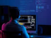 Person wearing headphones sits at a desk using a computer, with code displayed on the monitor and projected on the wall.