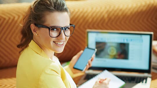 Woman wearing glasses and a yellow blazer holds a phone and notebook, sitting in front of an open laptop, smiling at the camera.