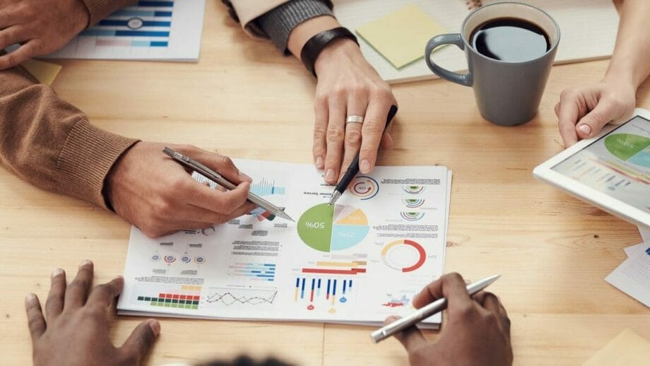 Four people discuss business charts and graphs on paper at a table with a cup of coffee and a tablet.