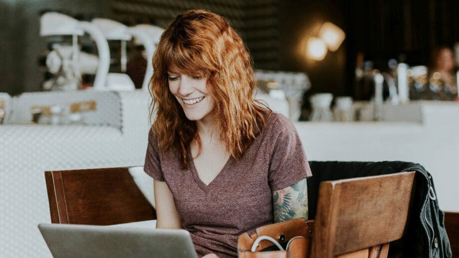 Woman with red hair sits in a café, smiling while looking at a laptop on a wooden table.