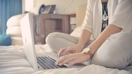 Person sitting cross-legged on a bed, typing on a laptop.