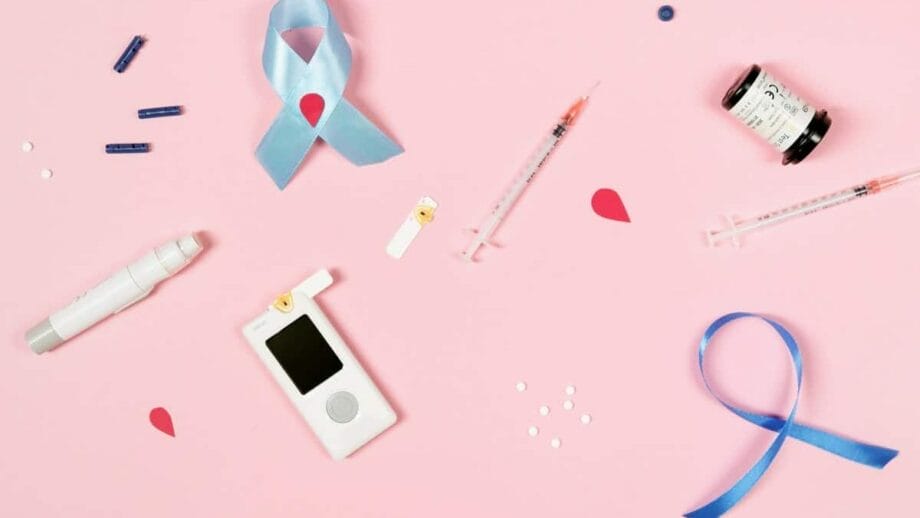A flat lay of diabetes management tools on a pink background, including a glucose meter, test strips, lancet devices, insulin syringes, and a vial. Blue awareness ribbons are also present.