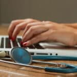 Hands typing on a laptop keyboard with a stethoscope lying on the wooden table nearby.