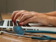 Hands typing on a laptop keyboard with a stethoscope lying on the wooden table nearby.