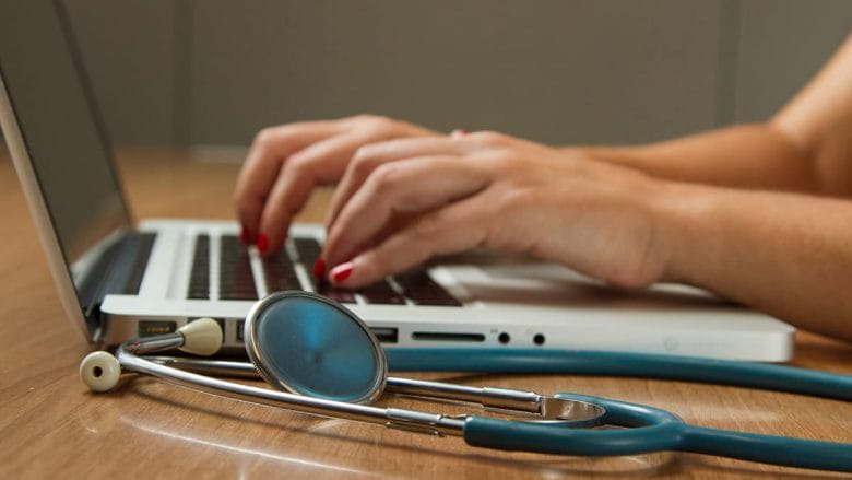 Hands typing on a laptop keyboard with a stethoscope resting on the wooden table nearby.