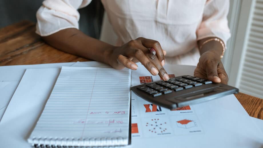 Person using a calculator at a wooden table with a lined notebook and various paperwork nearby.