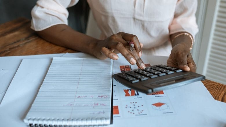 Person using a calculator at a wooden table with a lined notebook and various paperwork nearby.