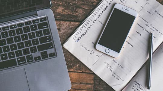 A laptop, smartphone, pen, and open planner with handwritten notes on a wooden desk.