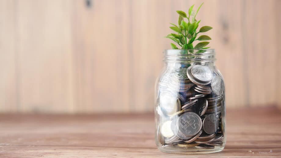 A glass jar filled with coins has a small green plant growing from the top, set on a wooden surface with a plain background.