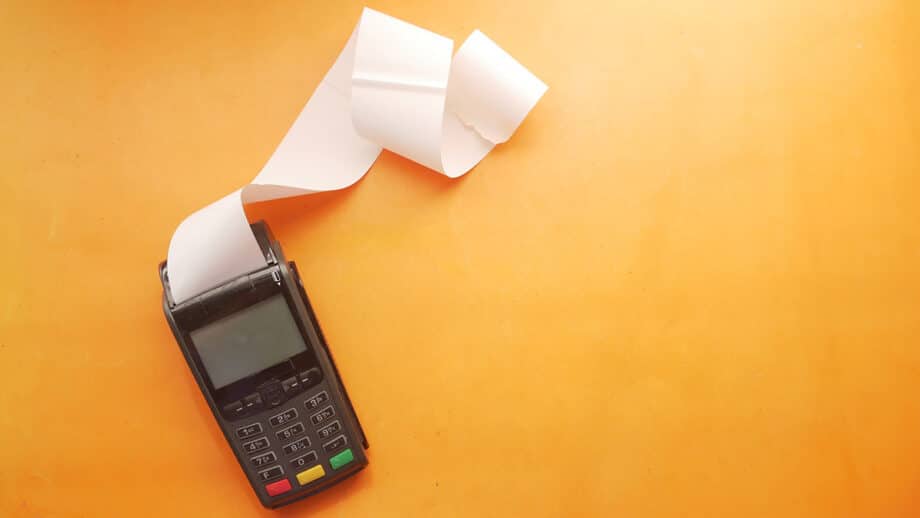A credit card payment terminal with a long receipt sits on an orange surface.