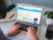 A person types on a MacBook laptop displaying an analytics dashboard. Nearby are a notebook, a plant, and a drink container.