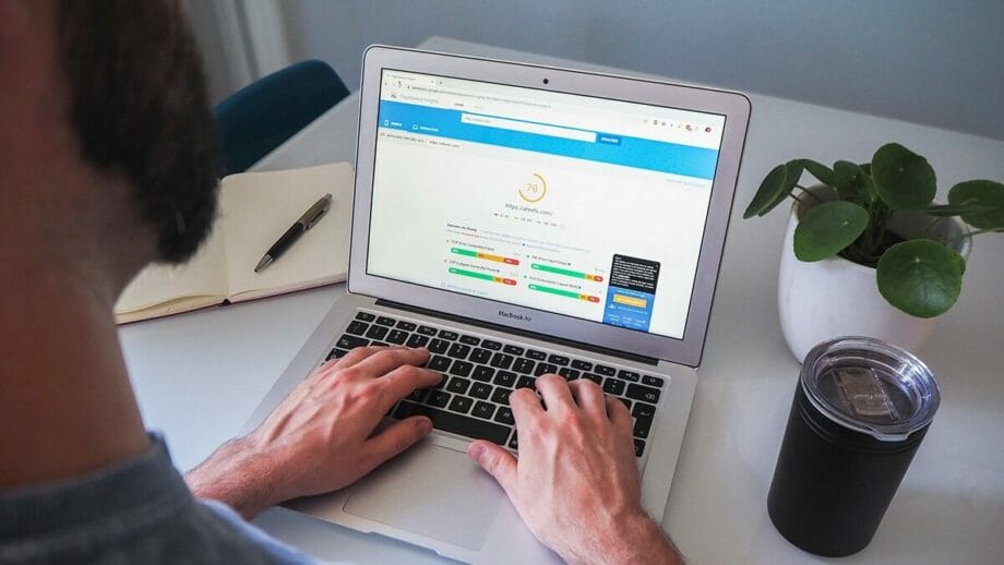 A person types on a MacBook laptop displaying an analytics dashboard. Nearby are a notebook, a plant, and a drink container.