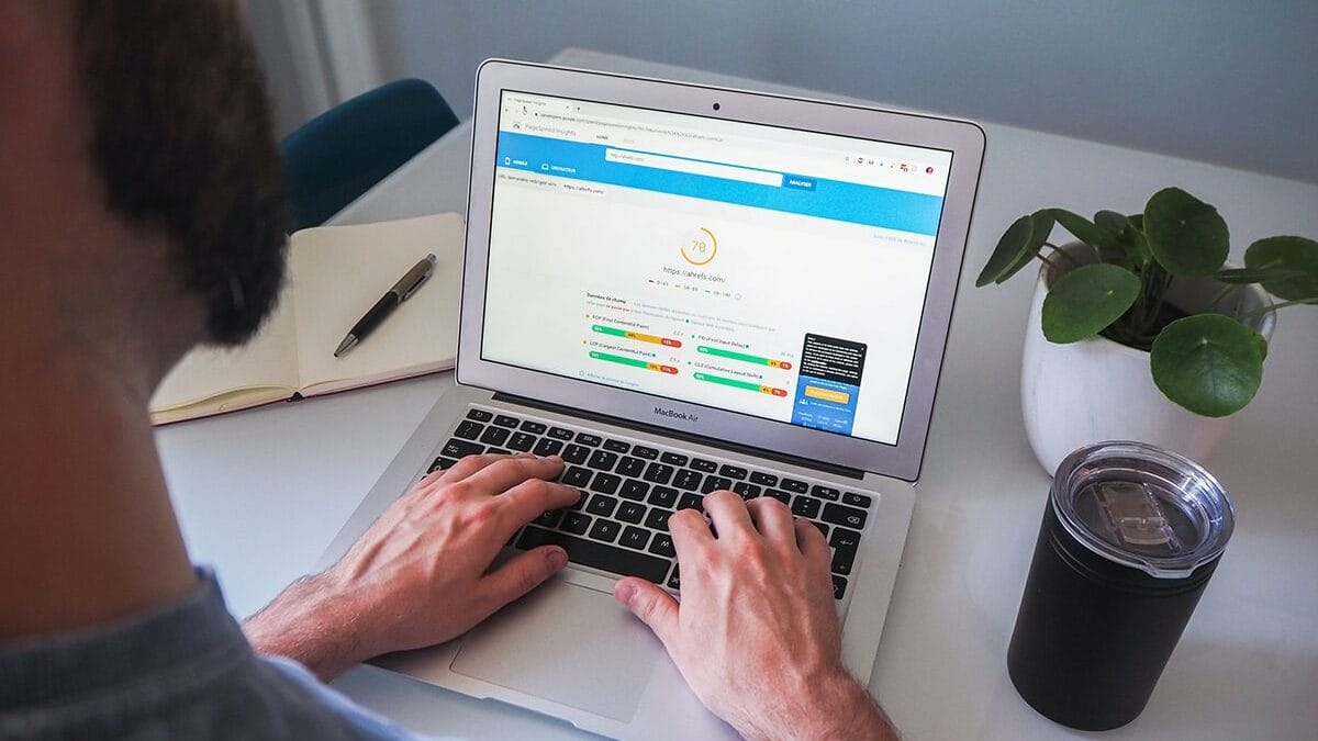 A person types on a MacBook laptop displaying an analytics dashboard. Nearby are a notebook, a plant, and a drink container.
