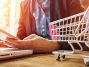 A person is holding a smartphone while sitting at a table with a laptop. A small toy shopping cart is placed on the table next to the laptop. The person is wearing a dark blazer over a blue shirt. The scene is brightly lit with a soft focus background.