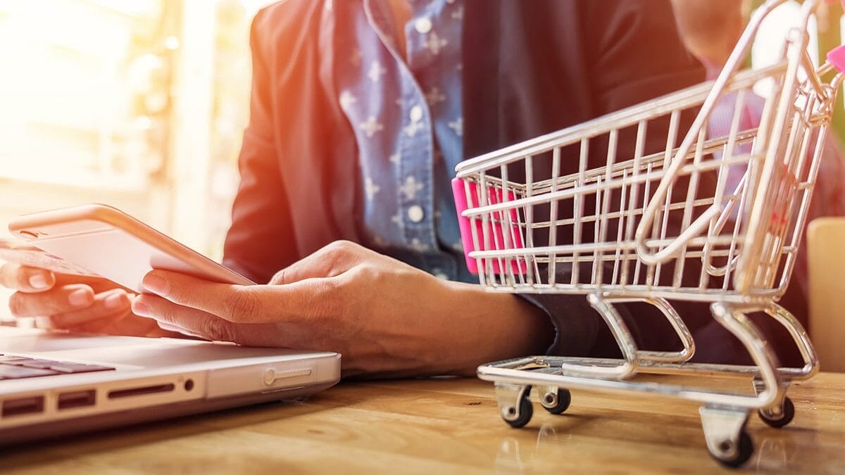 A person is holding a smartphone while sitting at a table with a laptop. A small toy shopping cart is placed on the table next to the laptop. The person is wearing a dark blazer over a blue shirt. The scene is brightly lit with a soft focus background.