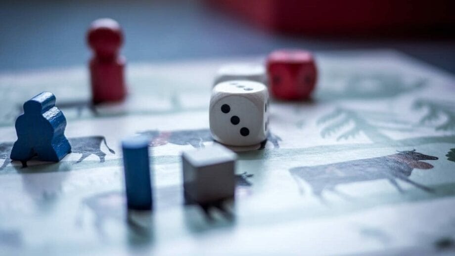 Close-up of a board game with focus on a white dice, surrounded by colorful meeple pieces on a detailed game board. the background features soft hues and light shadow play.
