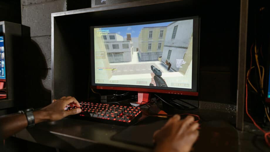 A person is playing a first-person shooter game on a desktop computer with a red-lit keyboard and mouse in a dark room. The screen displays a street scene in the game.