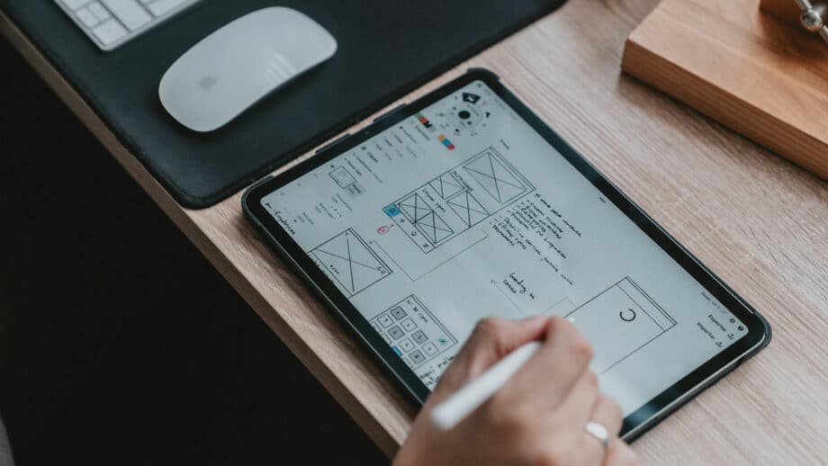 A person is using a stylus to work on a digital tablet that displays diagrams and notes, with a computer mouse and wooden desk accessories nearby.