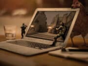 A chicken observes tiny soldiers engaged in a firefight on a laptop screen, with the scene depicted as if occurring within a rustic street setting. nearby are a mobile phone and a glass of water.