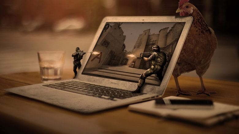 A chicken observes tiny soldiers engaged in a firefight on a laptop screen, with the scene depicted as if occurring within a rustic street setting. nearby are a mobile phone and a glass of water.