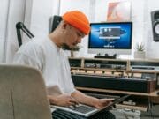 A person sits at a desk in a home studio, intently looking at a laptop, with audio equipment and a computer display in the background.