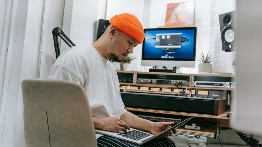 A person sits at a desk in a home studio, intently looking at a laptop, with audio equipment and a computer display in the background.