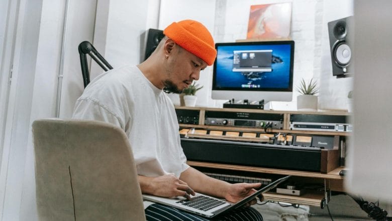 A person sits at a desk in a home studio, intently looking at a laptop, with audio equipment and a computer display in the background.