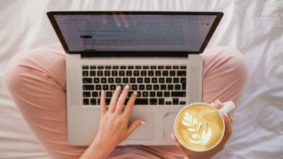 A person in pink pants sits cross-legged on a bed, working on a laptop while holding a cup of coffee with latte art.