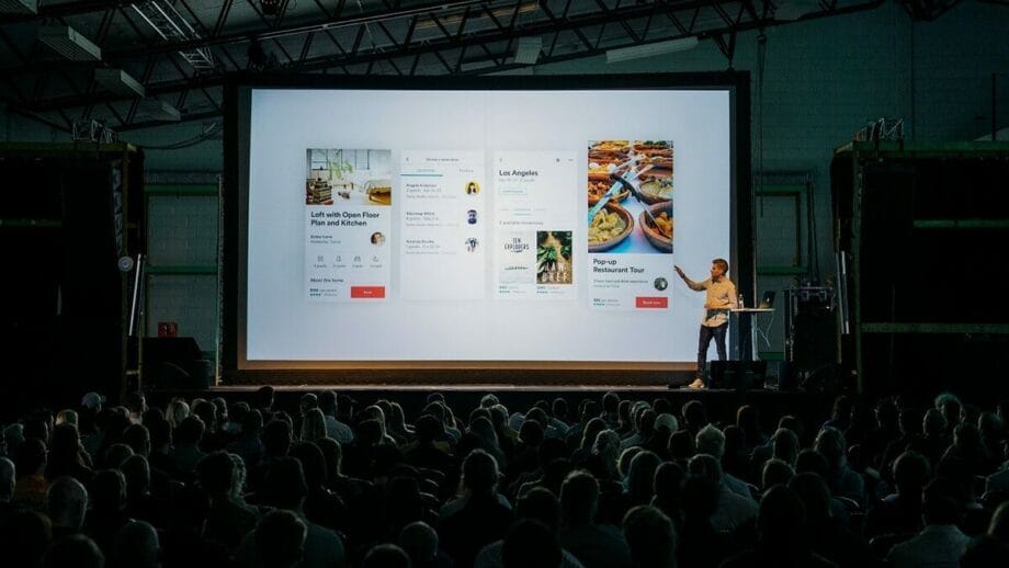 A speaker presents a slide show to a seated audience in a large, industrial-style auditorium.