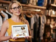 A woman in a yellow top and eyeglasses is holding a tablet with an online shop display on the screen, standing in a clothing store.