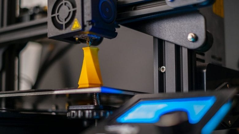 A 3D printer in operation, printing a yellow object on its platform. A control panel with blue and black display is visible in the foreground.