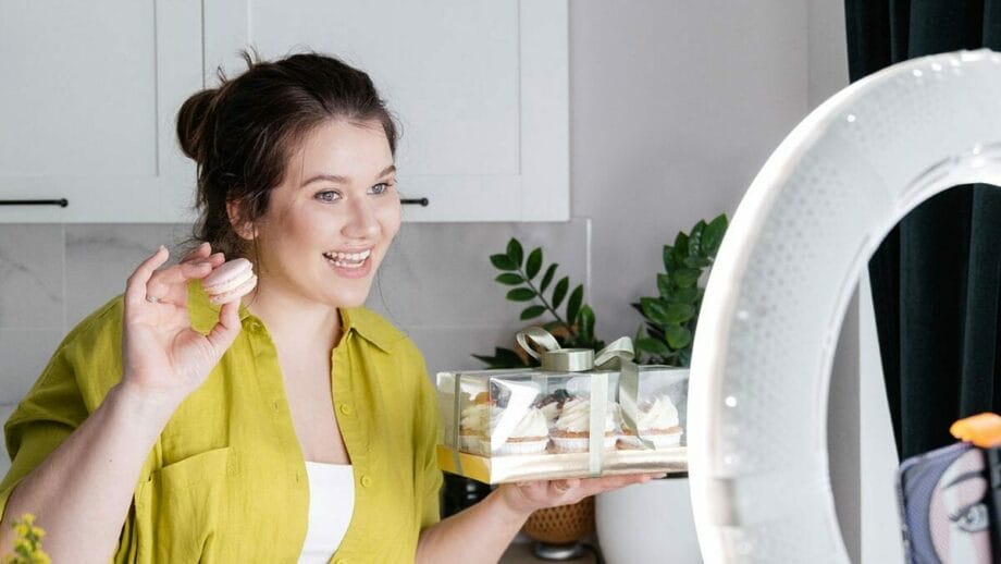 A woman in a green shirt holds a box of pastries and a single macaron, smiling in front of a ring light, likely recording a video or live stream in a kitchen setting.