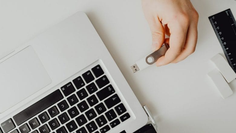 A hand inserts a USB flash drive into a laptop beside a black ruler and two white erasers on a white desk.