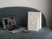 A laptop displaying an online shopping site sits on a gray couch next to two white shopping bags and a brown wallet.