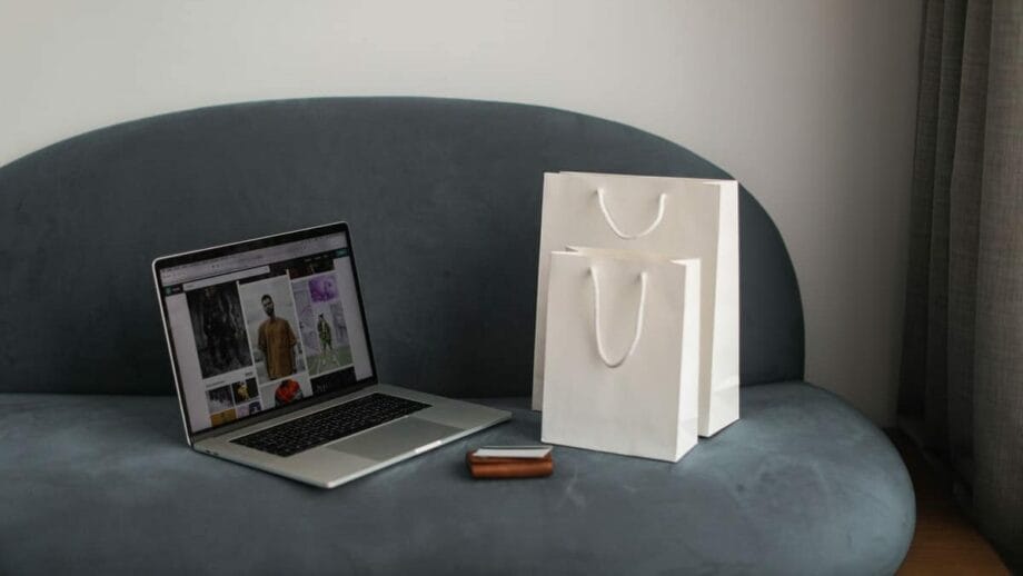 A laptop displaying an online shopping site sits on a gray couch next to two white shopping bags and a brown wallet.