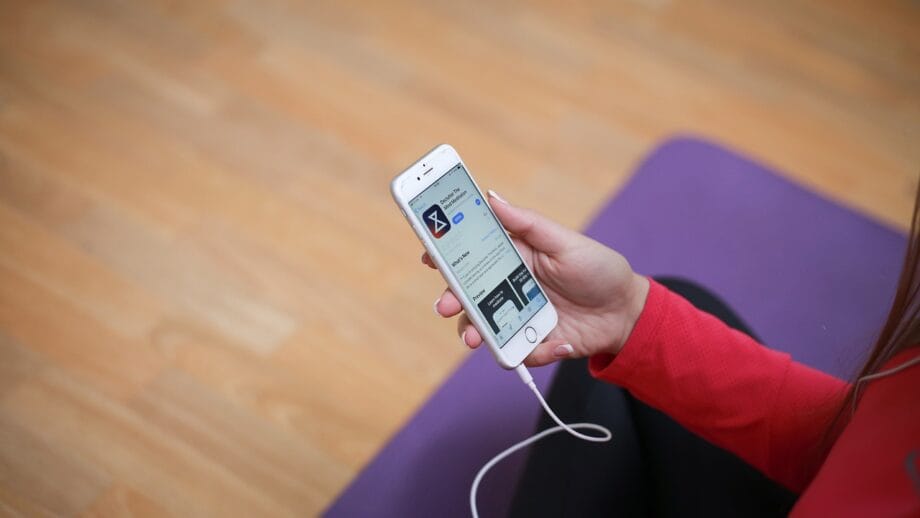 Person holding a smartphone displaying an app, with earphones plugged in. They are sitting on a purple cushion on a wooden floor.