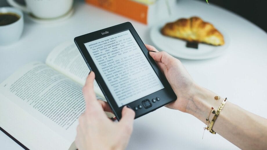 A person holding a kindle e-reader next to a physical book and a cup of coffee with a croissant on a white table.