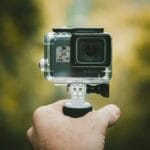 A hand holding a small action camera mounted on a handheld grip, with a blurred outdoor background.