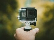 A hand holding a small action camera mounted on a handheld grip, with a blurred outdoor background.