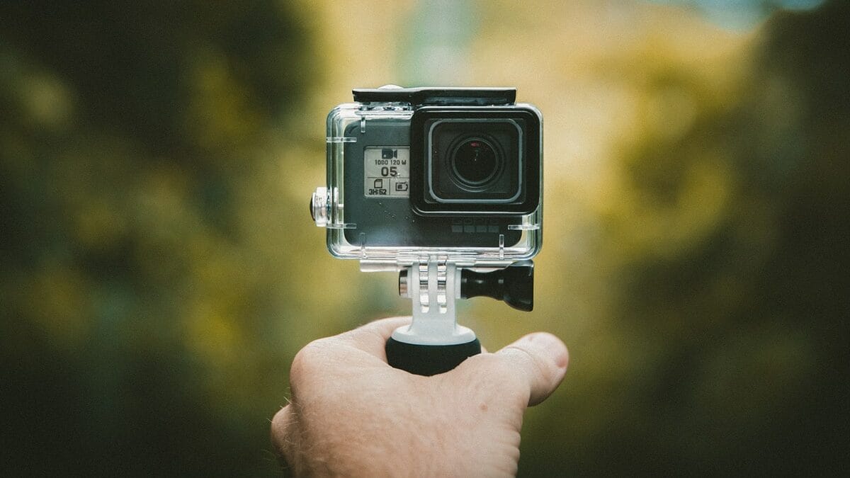 A hand holding a small action camera mounted on a handheld grip, with a blurred outdoor background.