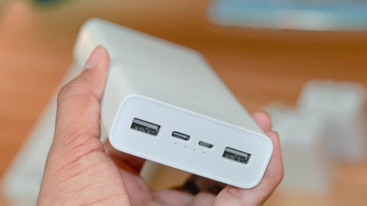 Hand holding a white power bank with two USB ports and one USB-C port, against a blurred background.