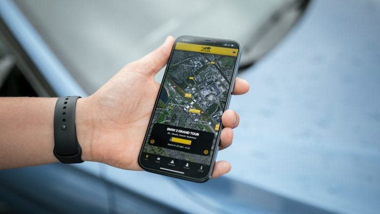 A hand holding a smartphone displaying a map with GPS locations on the screen, suggesting navigation or a tour guide app. The background features a blurred view of a car dashboard.