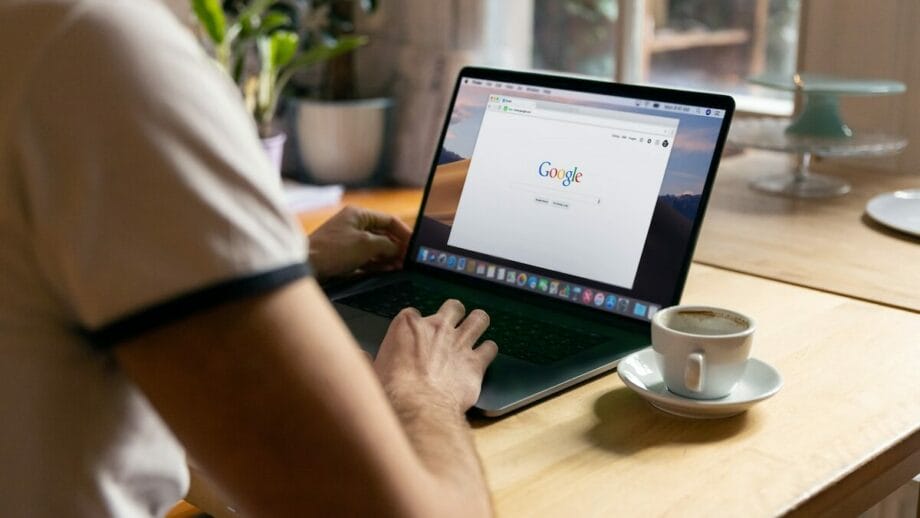 Person using a laptop on a wooden table with a Google search page open, a coffee cup nearby, and a plant in the background.