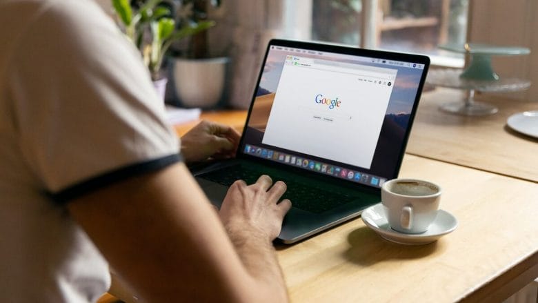Person using a laptop on a wooden table with a Google search page open, a coffee cup nearby, and a plant in the background.