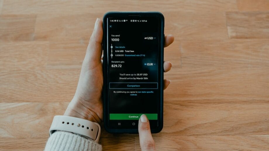 A person holds a smartphone displaying an online currency transfer screen, ready to confirm a payment.