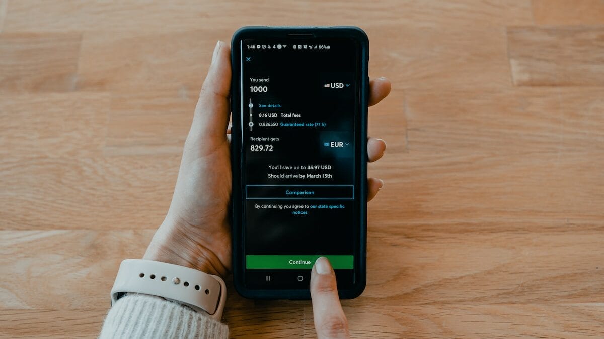 A person holds a smartphone displaying an online currency transfer screen, ready to confirm a payment.