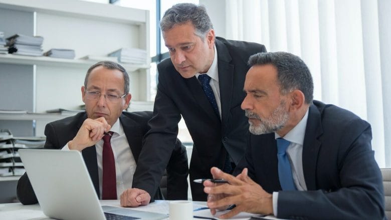 Three business professionals in suits are engaged in a discussion while looking at a laptop screen in an office environment.