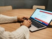 A person in a white sweater is typing on a laptop that is placed on a round wooden table. The laptop screen shows a web page with text. In the background, there are two beige chairs.