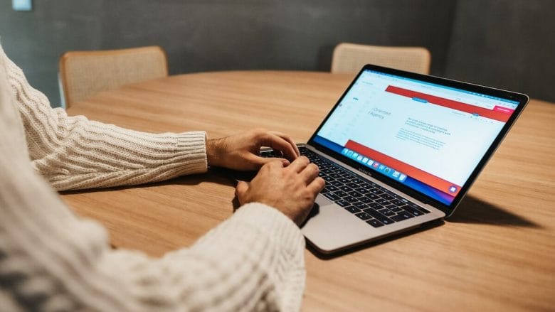 A person in a white sweater is typing on a laptop that is placed on a round wooden table. The laptop screen shows a web page with text. In the background, there are two beige chairs.
