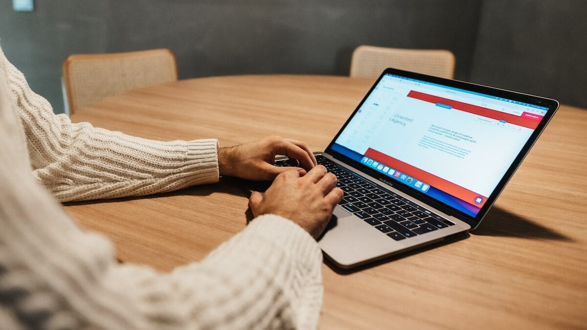 A person in a white sweater is typing on a laptop that is placed on a round wooden table. The laptop screen shows a web page with text. In the background, there are two beige chairs.